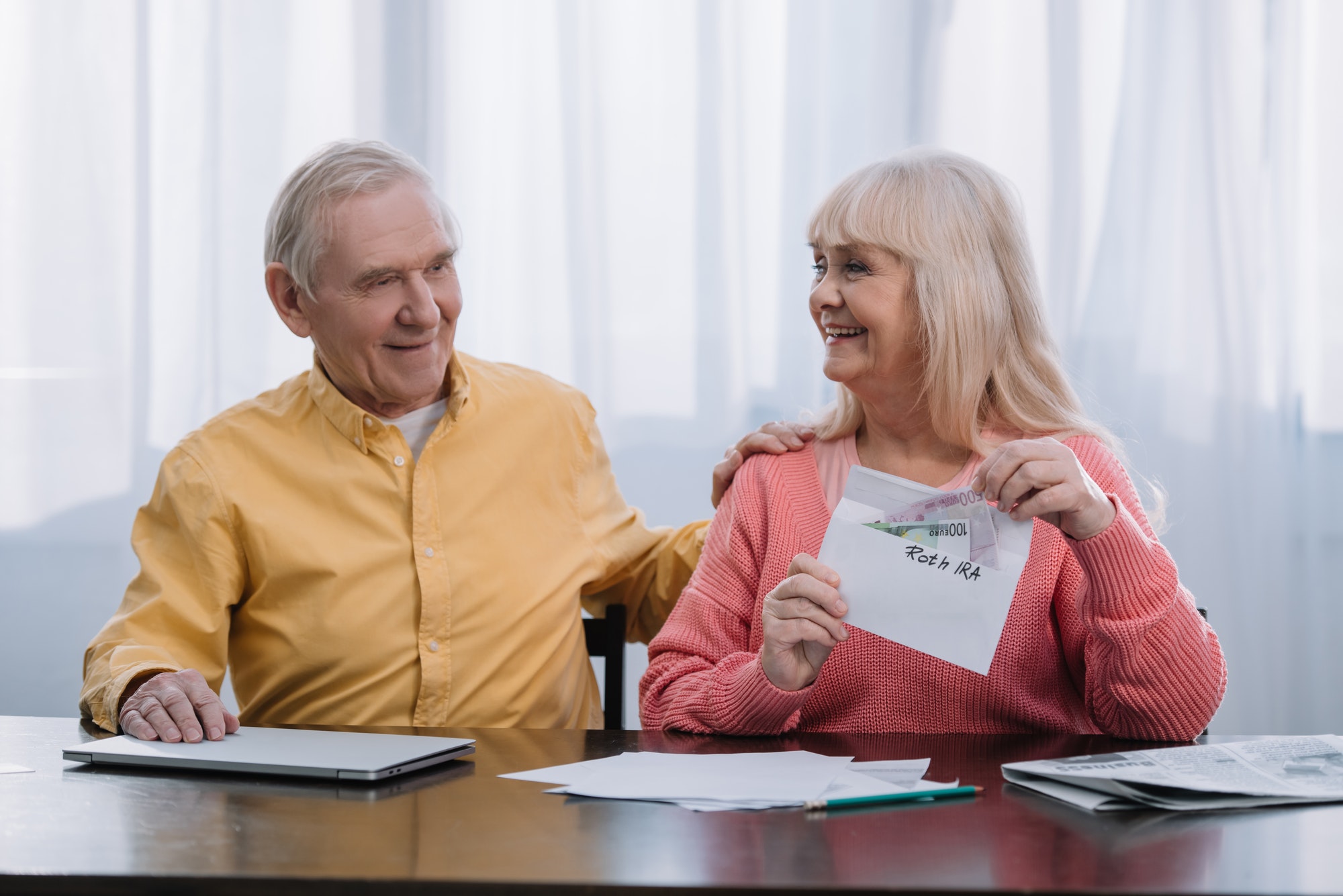 senior couple holding envelope with 'roth ira' lettering and money while sitting at table